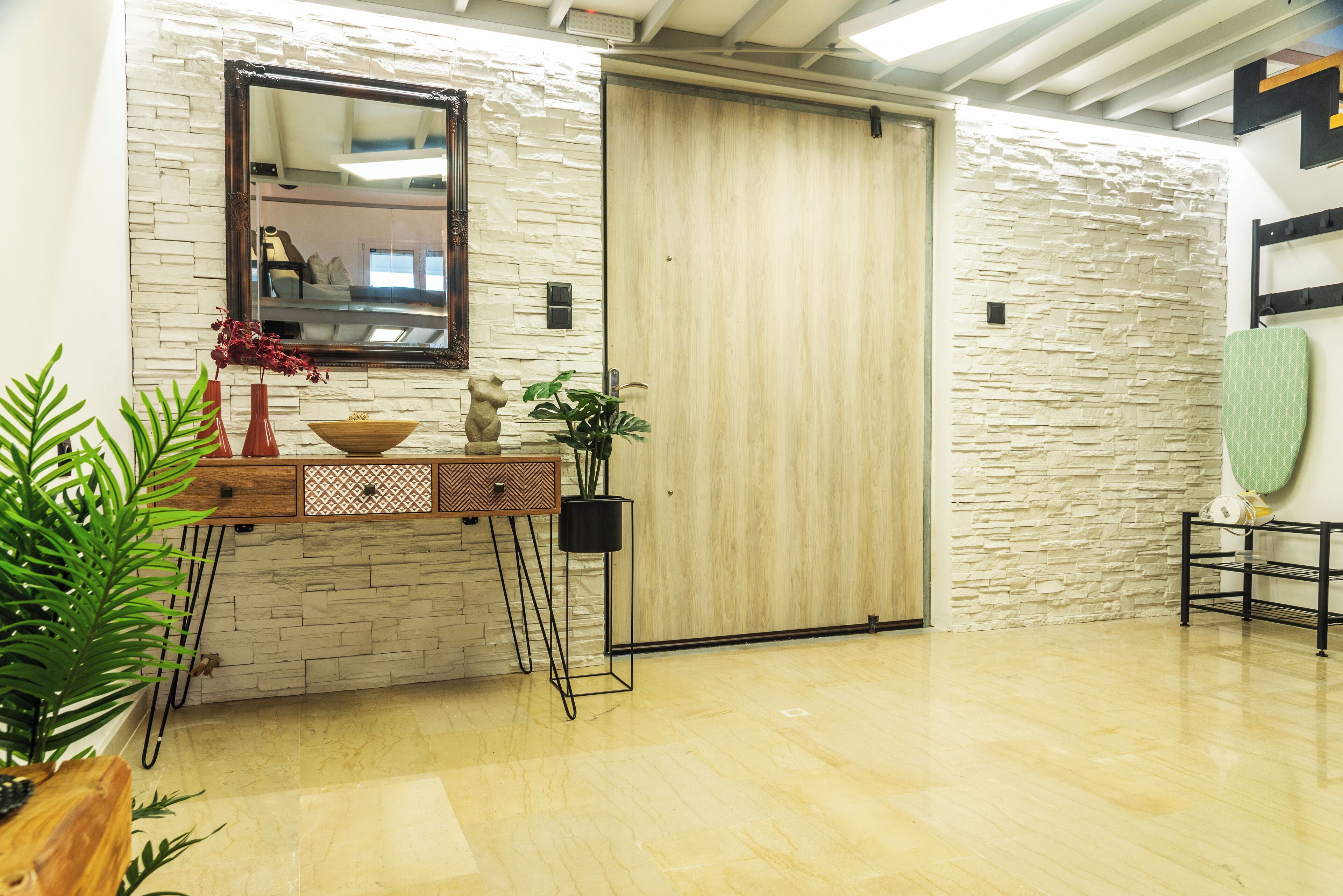 Una habitación con pared de piedra, puerta de madera, espejo, una consola con objetos decorativos y una planta en maceta.