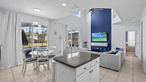 A modern kitchen with a central island, a dining area with white chairs, and a living room with a sofa and TV. - Ever After Adventure Villa (Kissimmee)