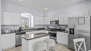A modern kitchen with white cabinetry, a central island with stools, stainless steel appliances, and a welcome sign on the fridge. - Ever After Adventure Villa (Kissimmee)