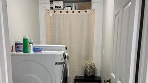 A laundry room with a front-loading washer and dryer, a small table with a vase, and a curtain. - BEAUTIFUL VACATION HOME          3 bedroom and 1 bathroom (Forest City)