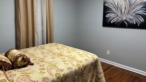 A bedroom with a bed, curtains, a floral bedspread, a wall-mounted painting, and wooden flooring. - BEAUTIFUL VACATION HOME          3 bedroom and 1 bathroom (Forest City)