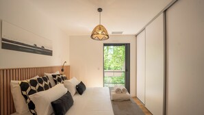 A bedroom with a bed, a hanging light fixture, a window with a view of greenery, and a door.