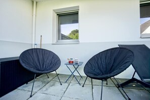 Un balcon avec deux chaises noires, une petite table sur laquelle se trouve un objet rouge, et une fenêtre donnant sur des arbres.