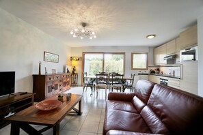 A living room with a brown leather sofa, a wooden coffee table, a TV, and a dining area with a table and chairs.