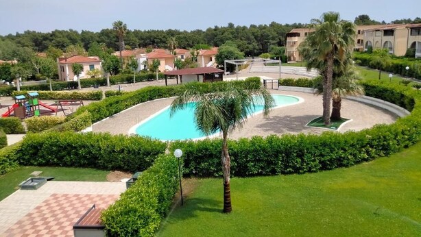 Piscina rodeada por um jardim bem cuidado, com uma palmeira e um espaço de estar com piso xadrez.