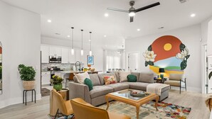 A modern living room with a sofa, coffee table, and chairs, a kitchen in the background, and a large wall mural. - Charleston-themed House for 10! (Goose Creek)