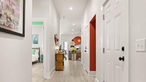A hallway with white walls, a wooden floor, and a door with a red accent. There are framed artworks on the walls and a plant on a side table. - Charleston-themed House for 10! (Goose Creek)