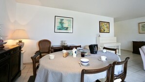 Een eetruimte met een ronde tafel, stoelen en diverse serviesartikelen. Er hangen ingelijste kunstwerken aan de muren.