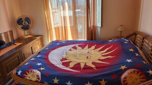 A room with a bed covered in a colorful blanket featuring a sun and stars, a wooden dresser, a desk with a computer, and a standing fan. - Casa Vacanze Relax Etna With Stunning Natural Beauty View (Giarre)