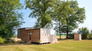 Una pequeña cabaña de madera con puerta roja, un cobertizo y un patio cubierto de césped y árboles.