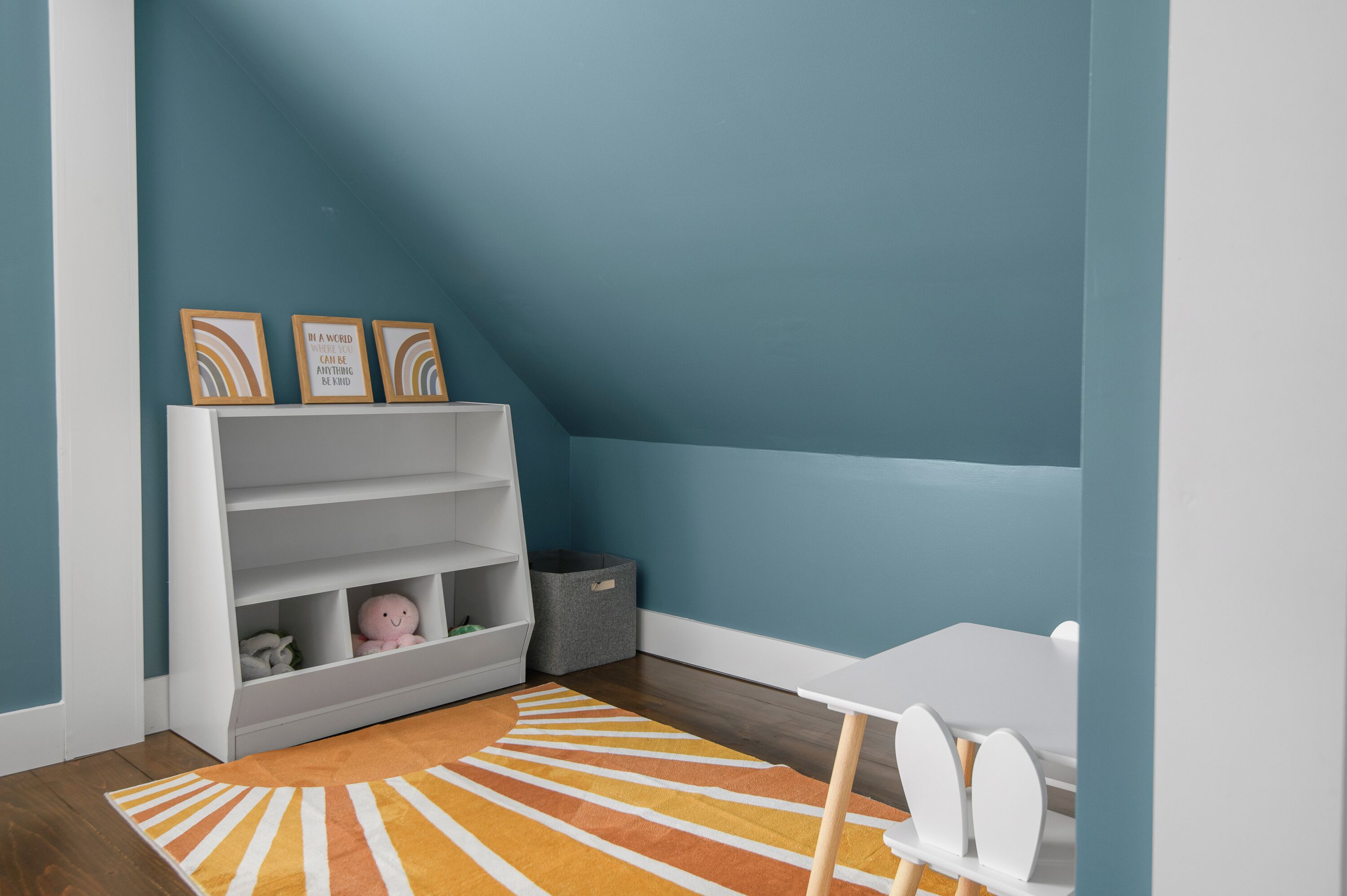 Une pièce d’angle avec une chaise blanche, une table blanche, une bibliothèque avec trois tableaux encadrés et un bac de rangement gris.