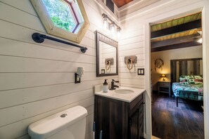 Ein Badezimmer mit Toilette, Waschbecken und Spiegel. Über dem Waschbecken befindet sich ein Fenster, und im angrenzenden Zimmer steht ein Bett.