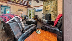 A cozy cabin interior with a stone wall, leather seating, a fireplace, and a large window overlooking a scenic view.