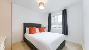 A neatly made bed with white bedding and two orange pillows, a bedside lamp, and a window with dark curtains. - Still Life Clerkenwell Deluxe (London)