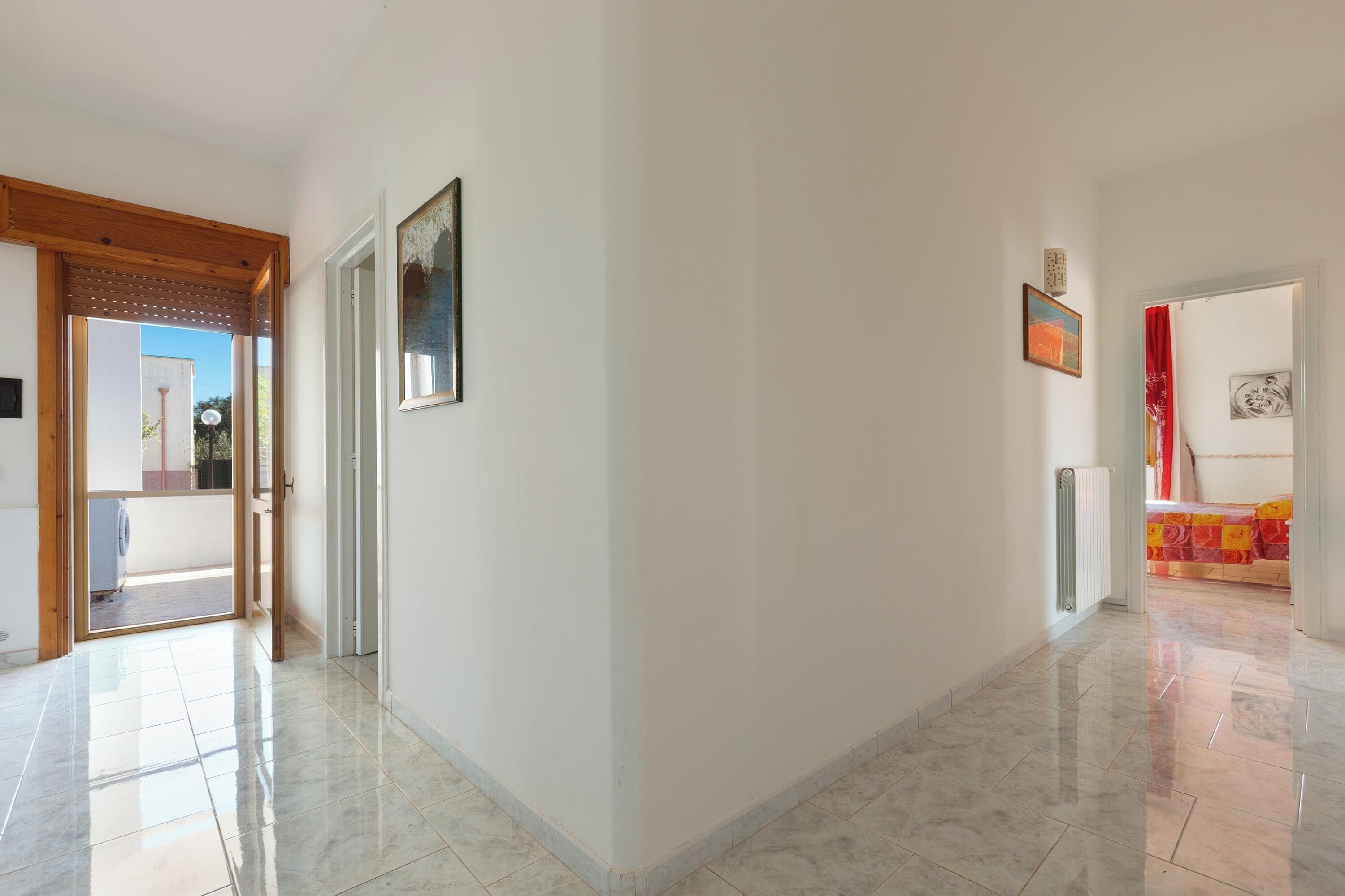 A hallway with a tiled floor, a wooden door frame, and a view into another room with a bed and curtains.