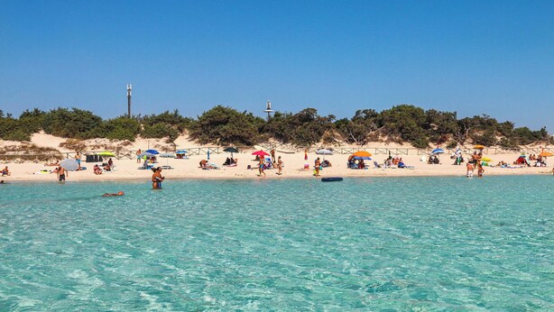 Ein Strand mit klarem türkisblauem Wasser, sandigem Ufer und Sonnenschirmen.