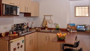 A compact kitchen with wooden cabinets, a microwave, a sink, and a small refrigerator. There are two black chairs and a bowl of fruit on the counter.