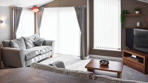 A modern living room with a grey sofa, a wooden coffee table, a flat-screen TV, and large windows with sheer curtains. - 2 bedroom accommodation in Moota, near Cockermouth (Moota, near Cockermouth)