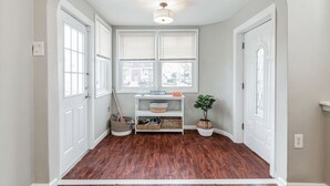 A hallway with wooden flooring, white walls, and a ceiling light fixture. There are two windows with blinds, a white door, and a small shelf with baskets and a plant.