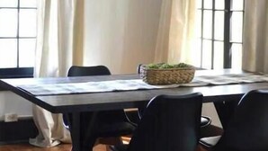 A dining area with a long table, black chairs, a chandelier, and large windows with curtains.