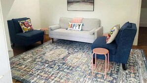 A living room with a grey sofa, two blue armchairs, a patterned rug, and a 'HAPPY' pillow. - The Backyard Bungalow (Batesville)