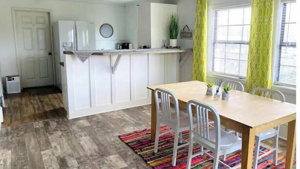 A compact kitchen with white cabinets, a wooden dining table with chairs, and a colorful rug. - The Backyard Bungalow (Batesville)