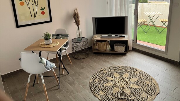 Een moderne woonkamer met een flatscreen televisie, een eettafel met stoelen, een groot geweven vloerkleed en uitzicht op een groene buitenruimte door een schuifdeur van glas.