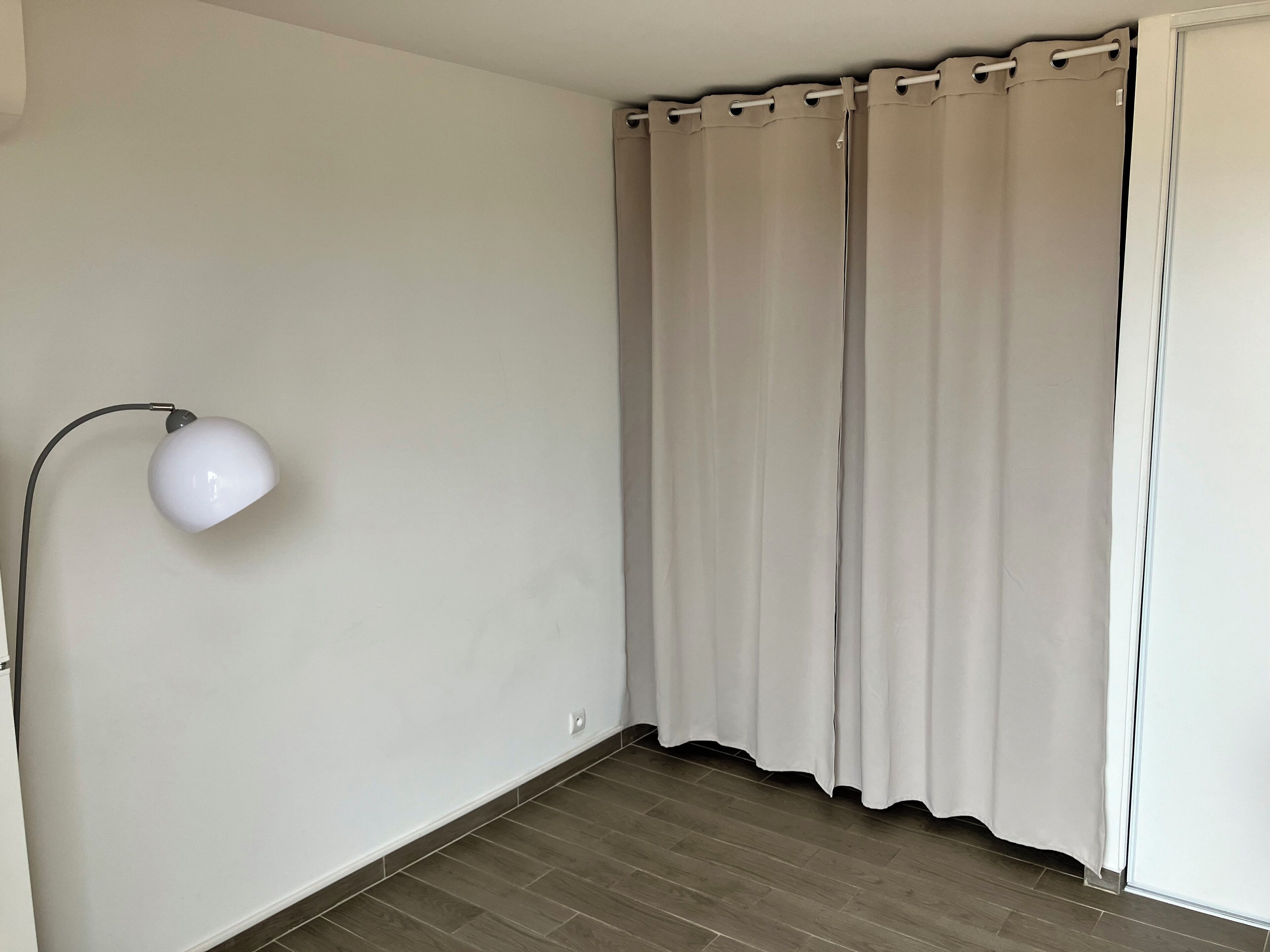 Une pièce avec un mur blanc, un lampadaire et une fenêtre ornée de rideaux beiges.