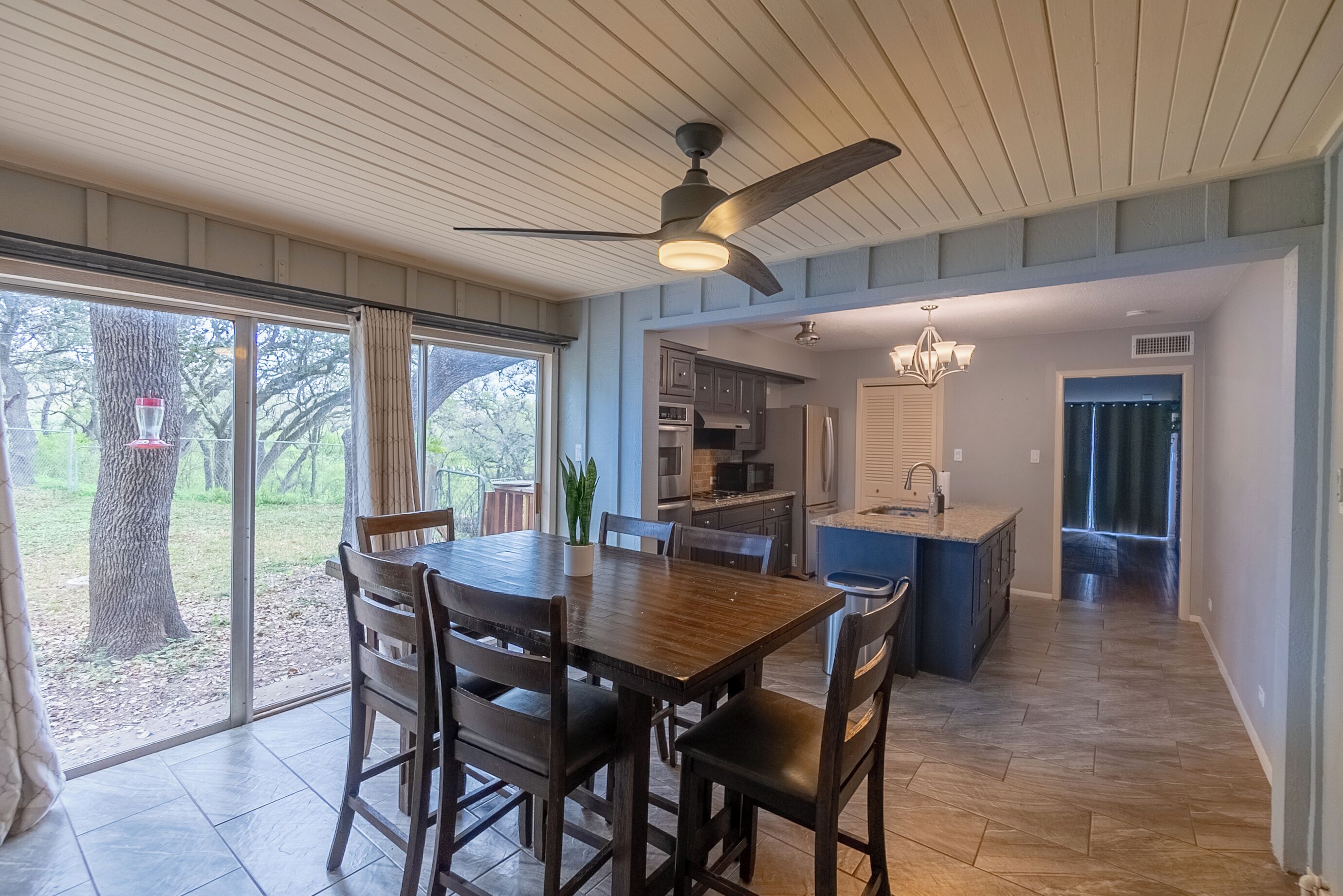 A spacious dining area with a wooden table and chairs, a ceiling fan, and large windows offering a view of the outdoors.