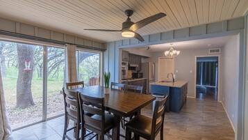 A spacious dining area with a wooden table and chairs, a ceiling fan, and large windows offering a view of the outdoors.