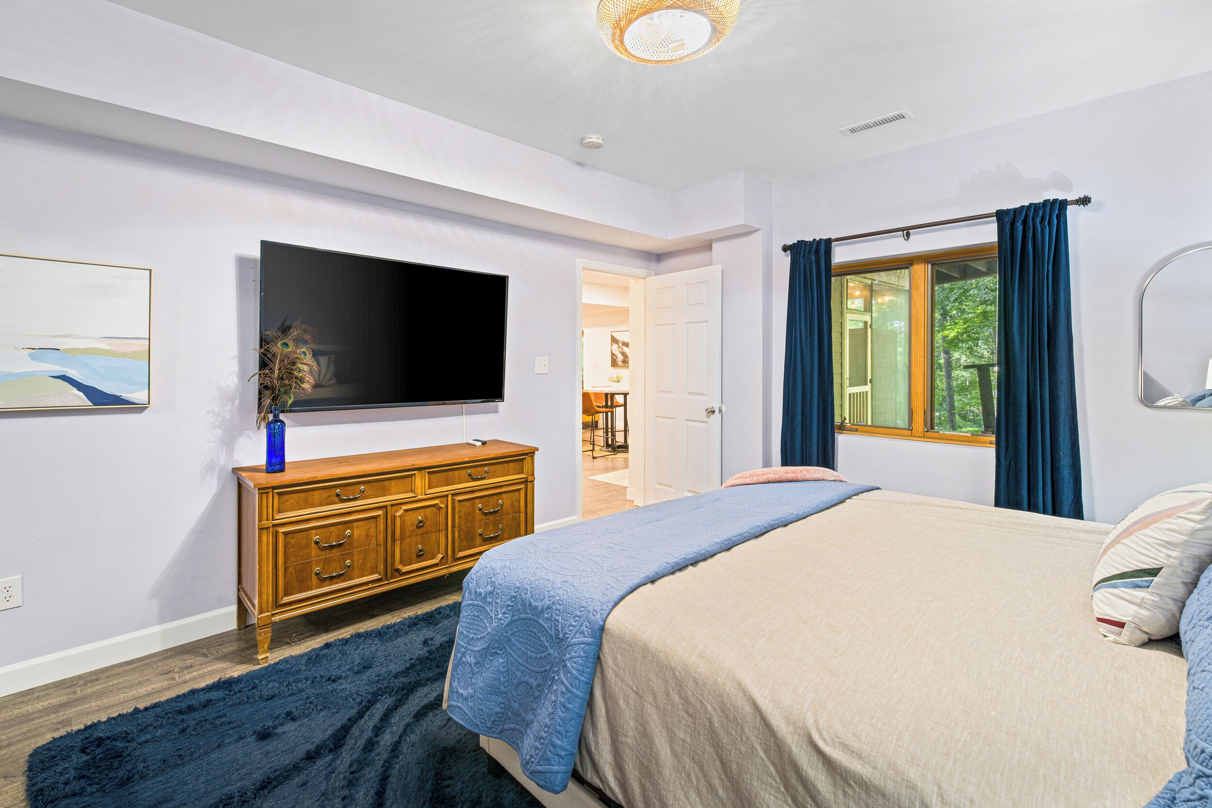 A bedroom with a bed, a dresser, a flat-screen TV, a window with curtains, and a painting on the wall.