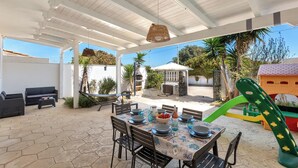An outdoor patio with a dining table set for four, surrounded by chairs, and a children's play area in the background.