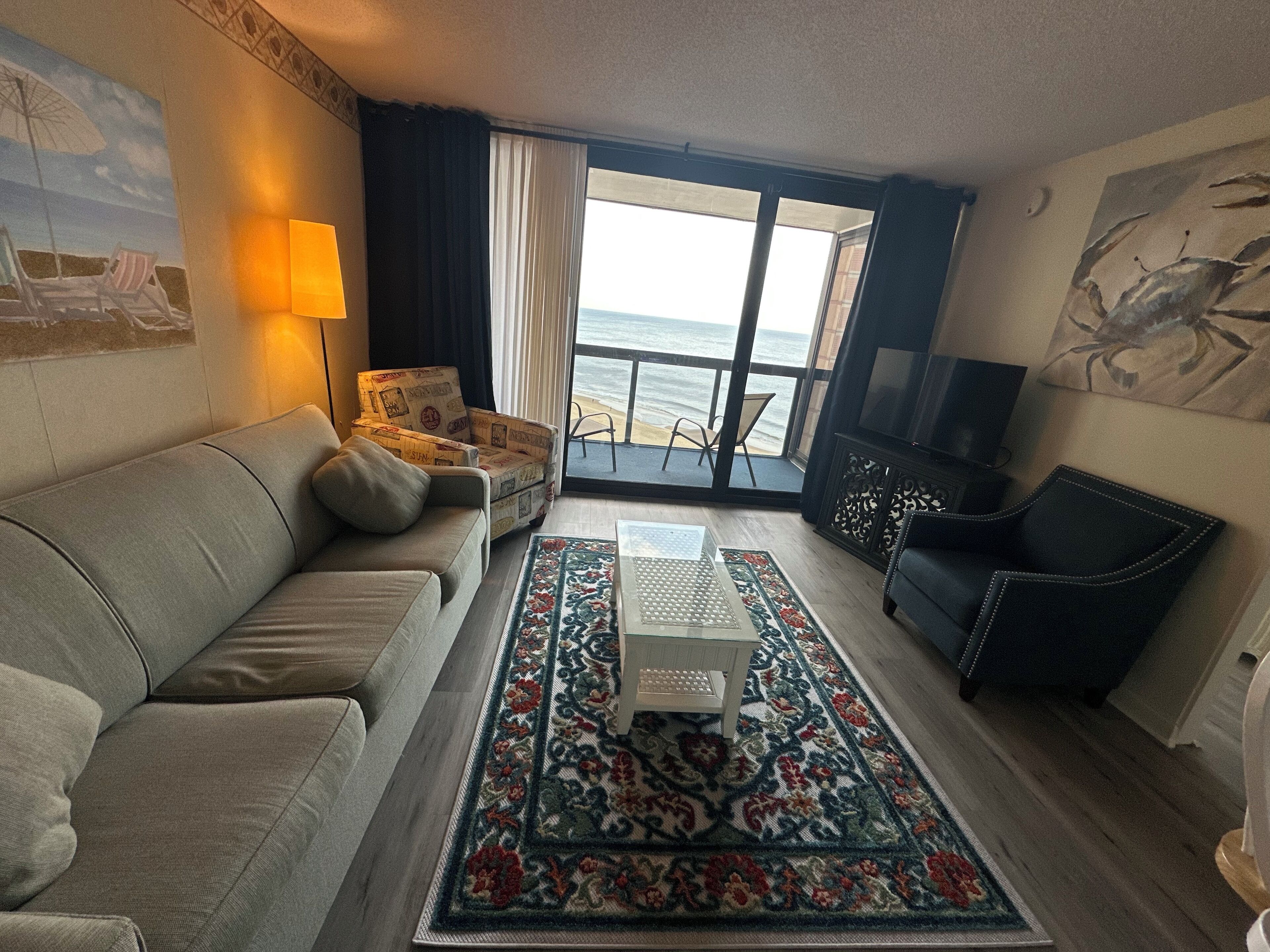 A living room with a sofa, a coffee table, a patterned rug, a television, and a view of the ocean through large windows.