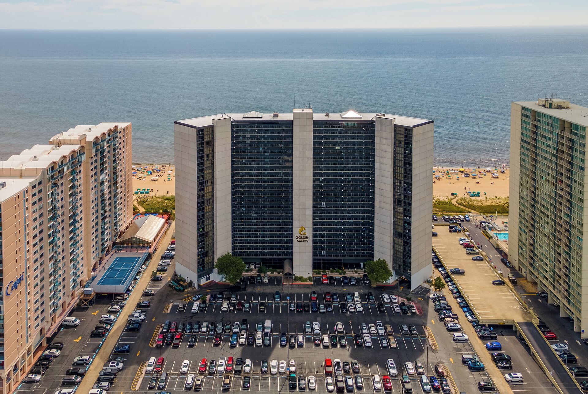 A high-rise hotel with a parking lot in front, overlooking a beach and the ocean.