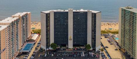 A high-rise hotel with a parking lot in front, overlooking a beach and the ocean.
