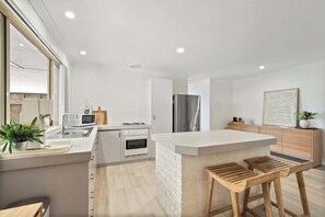 A modern kitchen with white cabinetry, a stainless steel refrigerator, wooden stools, and a white island.