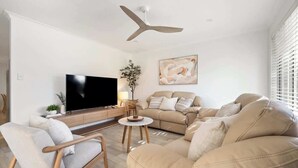 A living room with a sofa, armchair, TV, ceiling fan, and artwork on the wall. - Luna Getaway—Spacious Family Escape by the Coast (Quindalup)