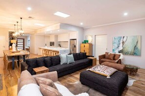 A modern living room with a sofa, ottoman, and coffee table, a dining area with a table and chairs, and a kitchen in the background.