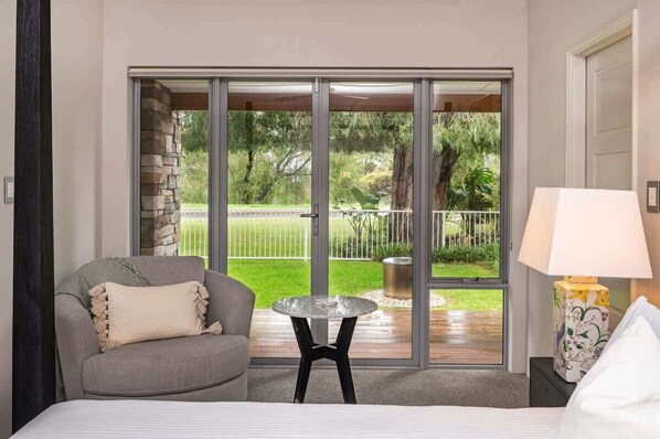 A hotel room with a bed, a gray armchair, a small round table, a lamp, and a sliding glass door leading to a balcony with a view of greenery.