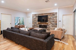 A living room with a stone fireplace, a large flat-screen TV, a dark-colored sofa, and a light-colored armchair.