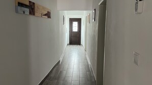 Een smalle gang met een deur aan het einde, een intercom bevestigd aan de muur en een lijst met een afbeelding aan de linkerwand.