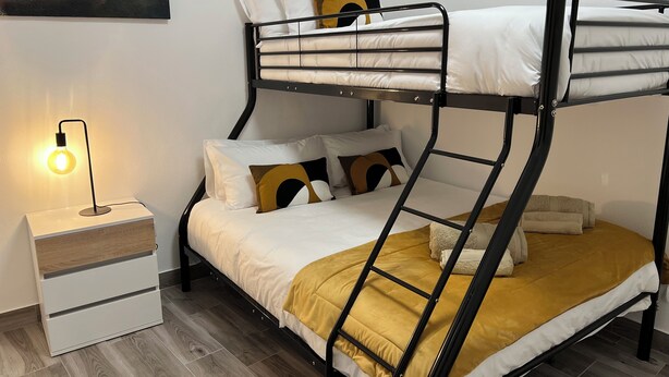 Een stapelbed met een ladder, wit beddengoed en een mosterdgele plaid. Er staat een nachtkastje met een lamp en een lade.