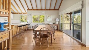 A spacious dining area with a long wooden table and chairs, a kitchen in the background, and large windows offering a view of the outdoors. - Mariners Rest—Bushland Haven w/ Stunning Sea Views (Eagle Bay)