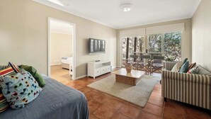 A spacious living room with a sofa, a television, a dining area, and a large window. - Mariners Rest—Bushland Haven w/ Stunning Sea Views (Eagle Bay)