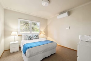 A hotel room with a bed, two bedside lamps, a window with blinds, a white bedside table, and a white dresser.