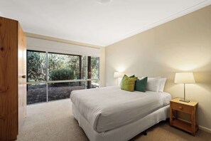 A bedroom with a large window, a bed with pillows, a bedside table with a lamp, and a wooden door.