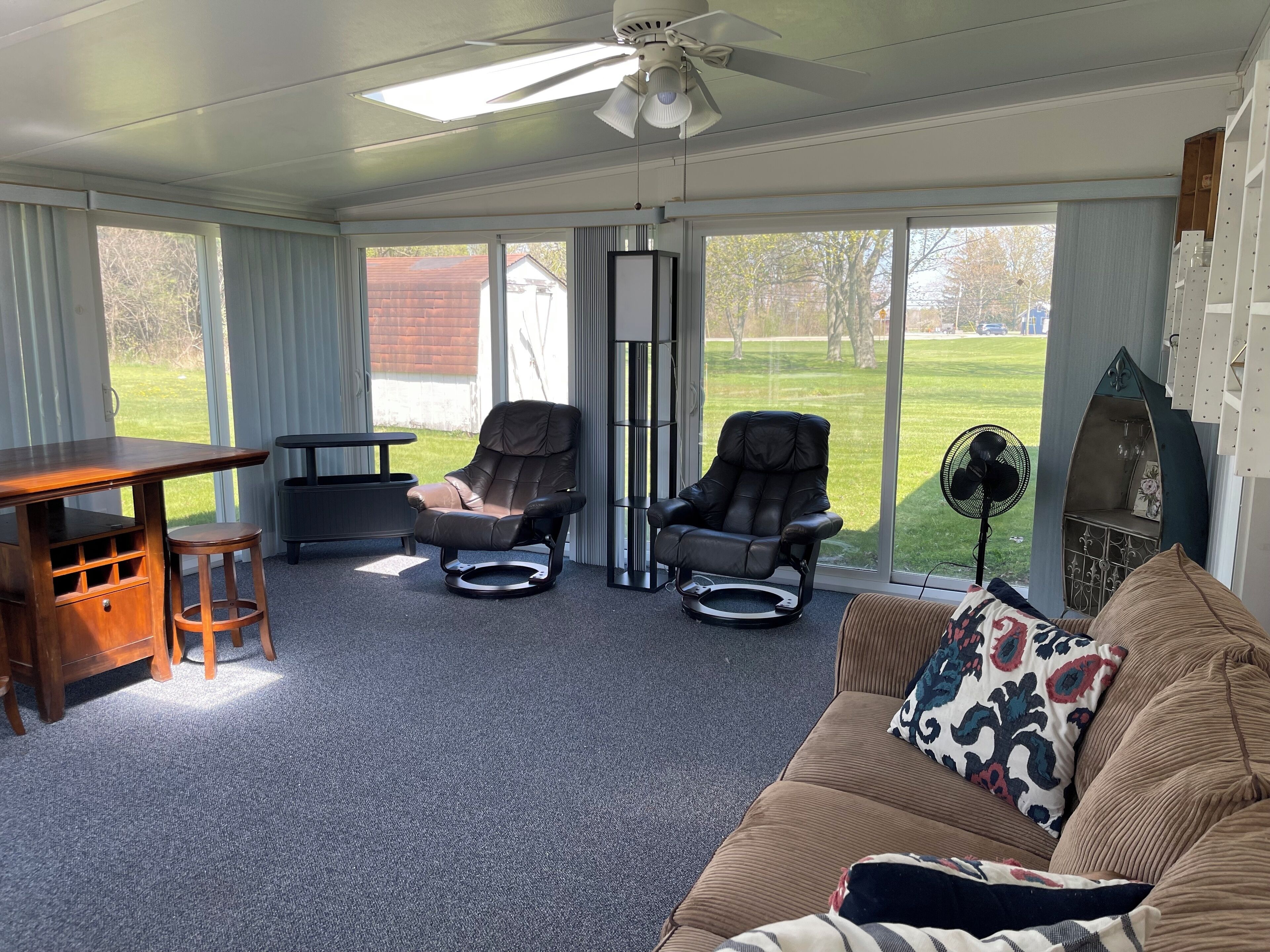A sunlit room with a ceiling fan, two reclining chairs, a wooden table, a couch with patterned pillows, and a large window offering a view of a grassy yard.