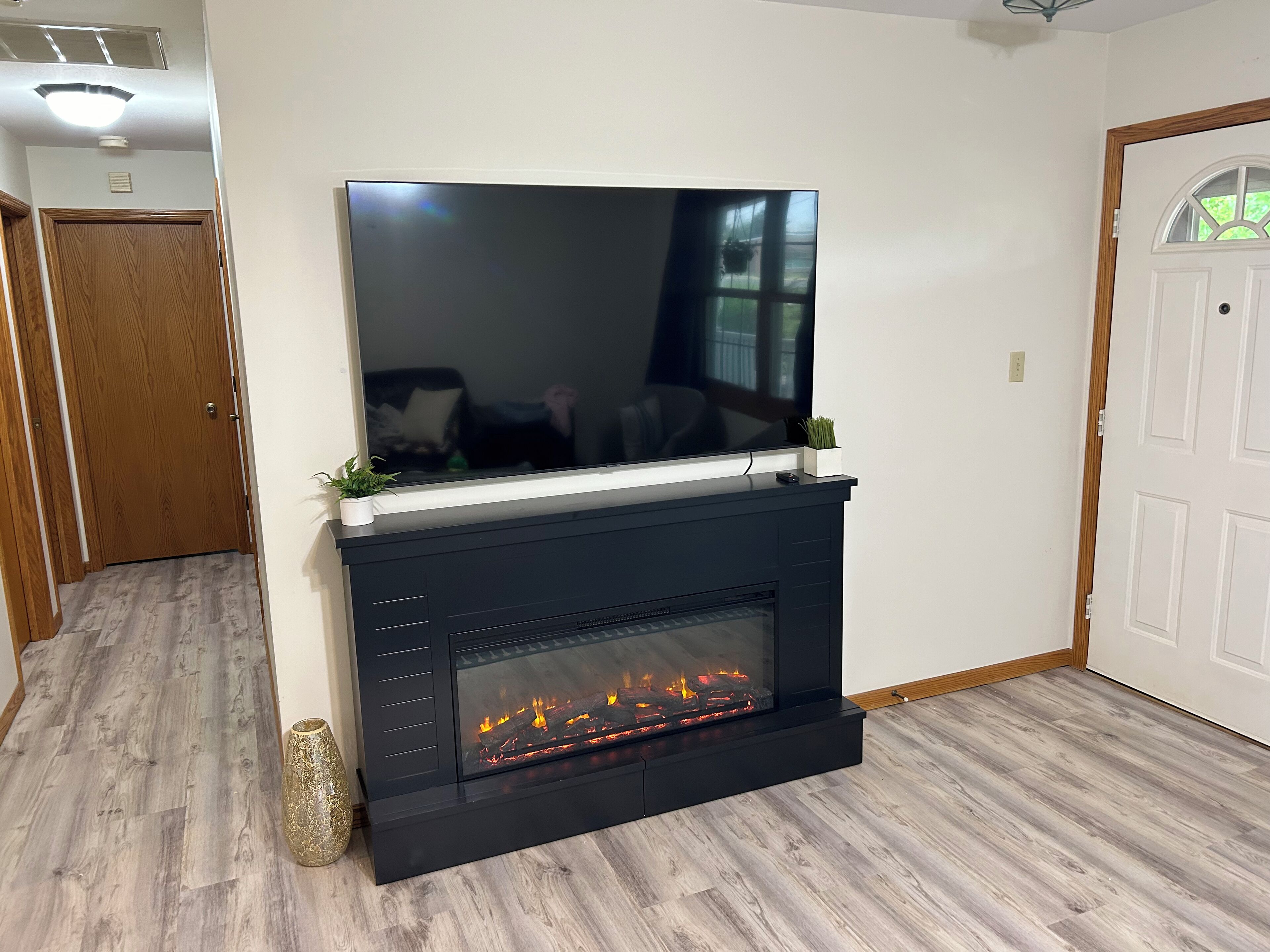 A modern living room with a flat-screen TV mounted above a black fireplace, wooden flooring, and a white door.