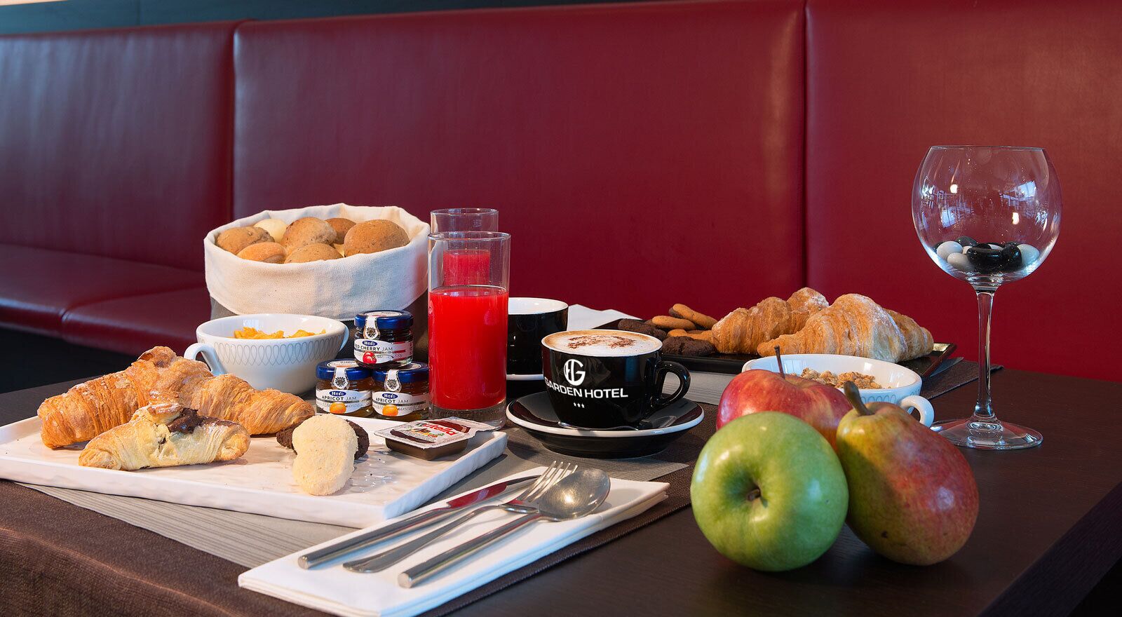 Desayuno con cruasanes, manzanas, un vaso de jugo de frutas rojas y una taza de café con la etiqueta 'Garden Hotel'.