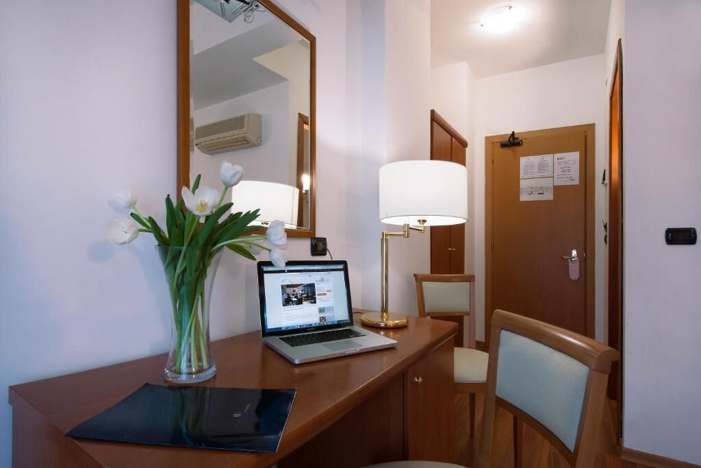 Una habitación de hotel con un escritorio de madera, una computadora portátil, un florero con flores, una lámpara y una silla.